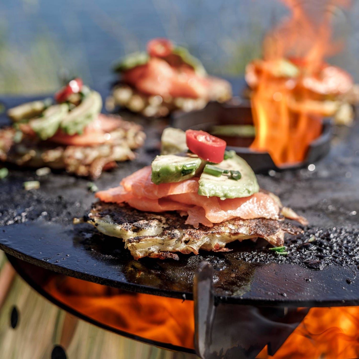 Close-up of juicy grilled salmon topped with avocado slices, chives, and a red chili pepper on a handcrafted steel griddle with open flame, ideal for outdoor cooking in camping and grilling collections.