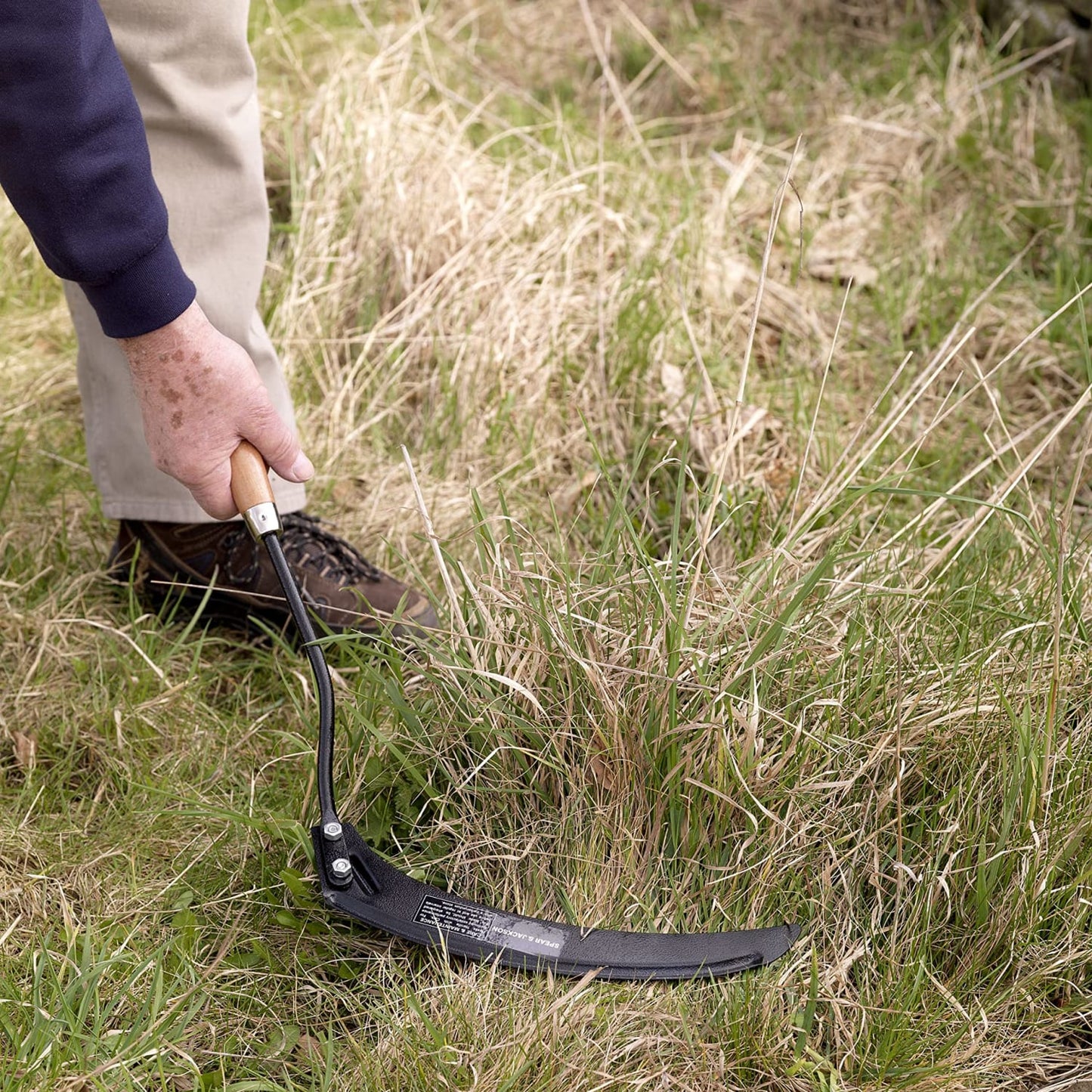 Handheld grass hook with carbon steel black hammer powder-coated blade and weatherproofed hardwood handle cutting tall dry grass, featured in Farm, Garden & Tool and Lawncare collections.