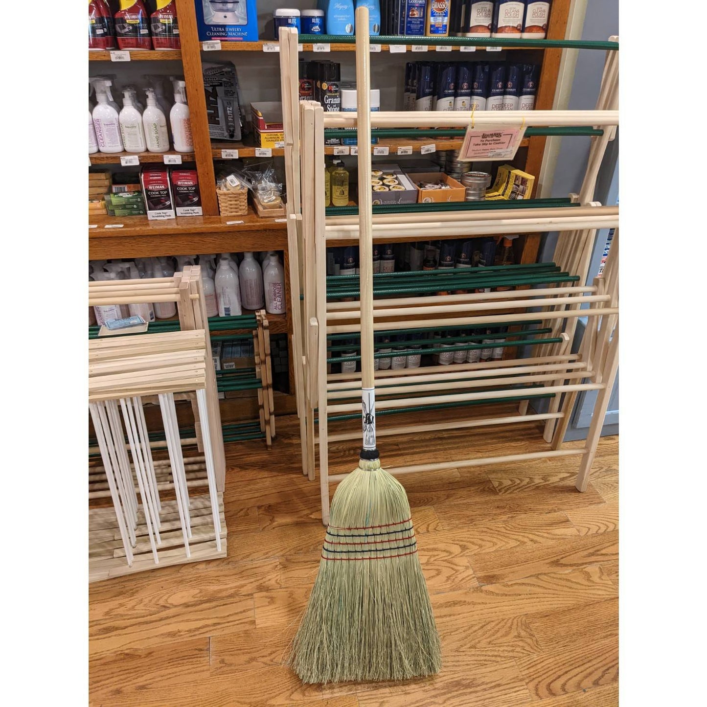 Handcrafted Amish made large corn broom with natural straw bristles bound by multiple rows of stitching, standing on a wooden floor near household and cleaning supplies, USA made.