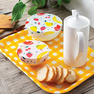 Two colorful reusable bowl covers with elastic bands securing fruit-printed plastic covers over bowls, placed on a yellow checkered tray with white ceramic teapot and sliced bread, featured in Kitchen & Food, Storage and Serving, Best Sellers, and Promo Eligible collections.