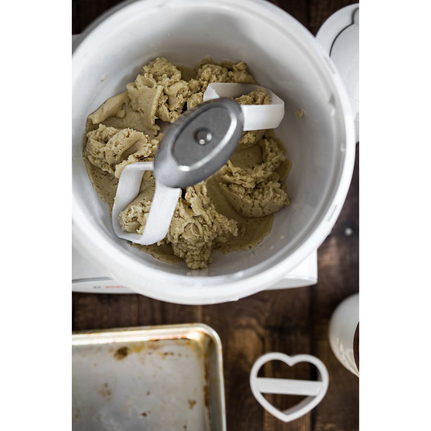 Close-up of thick cookie dough being mixed in a white bowl by dual cookie paddle attachments, accompanied by a heart-shaped cookie cutter on a wooden surface, USA Made kitchen gadget.