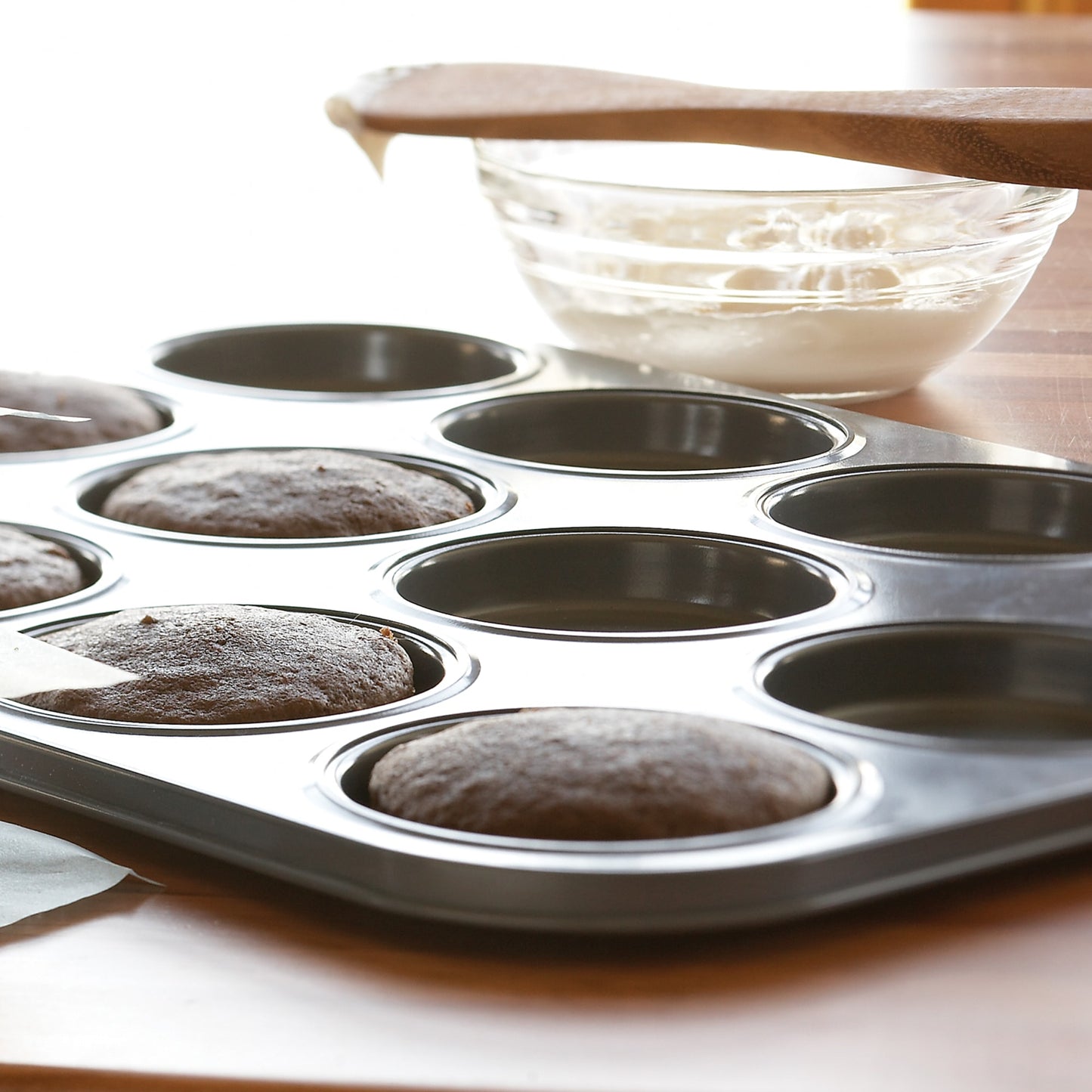Carbon steel whoopie pie pan with six freshly baked chocolate cakes rising evenly in shallow cavities, a glass bowl of white frosting and wooden spatula resting nearby, ideal for Kitchen & Food, Cookware, Baking Supplies, Best Sellers, and Promo Eligible collections.