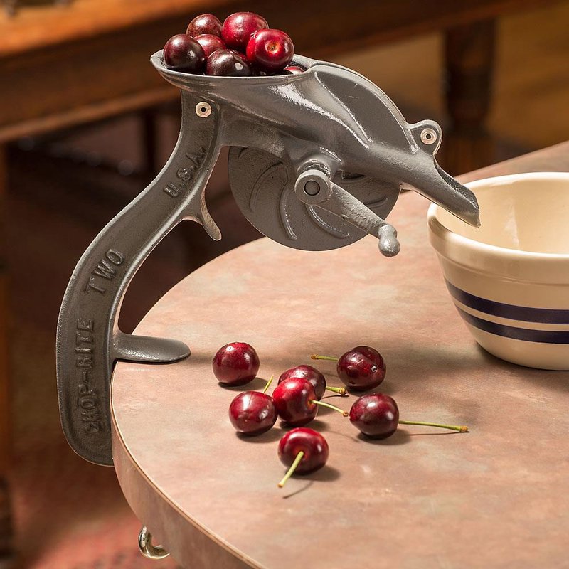 Handcrafted antique-style cherry pitter made in USA with durable cast body and crank wheel, shown with fresh cherries on a kitchen table, ideal for Kitchen & Food and Specialty Equipment collections.