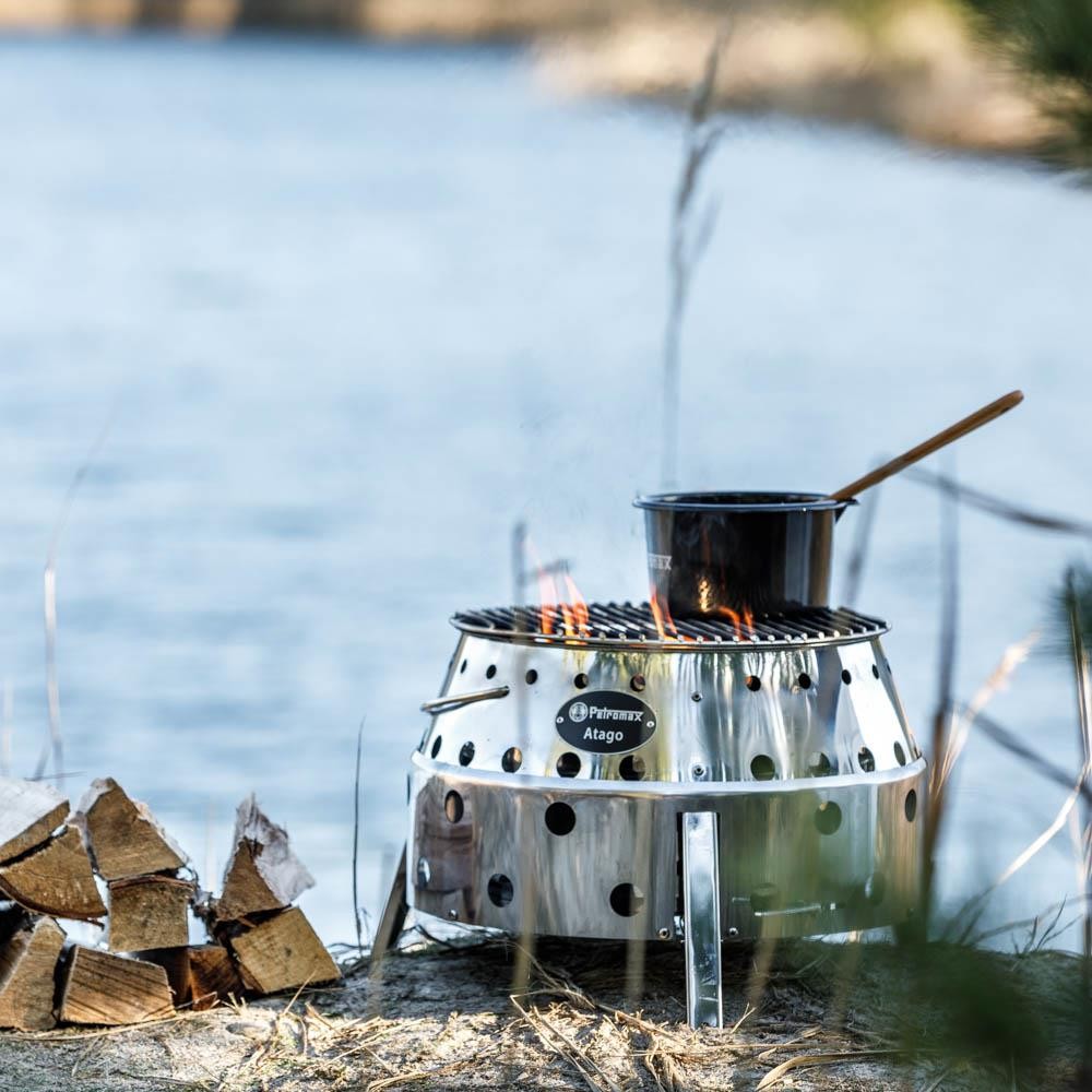 Black enamel pan with a wooden handle simmering over an open flame on a stainless steel Petromax Atago stove near a lakeshore, ideal for camping and outdoor cooking in Farm, Garden & Tool, Kitchen & Food, Camping, Cookware, Pots and Pans, Best Sellers, and Promo Eligible collections.