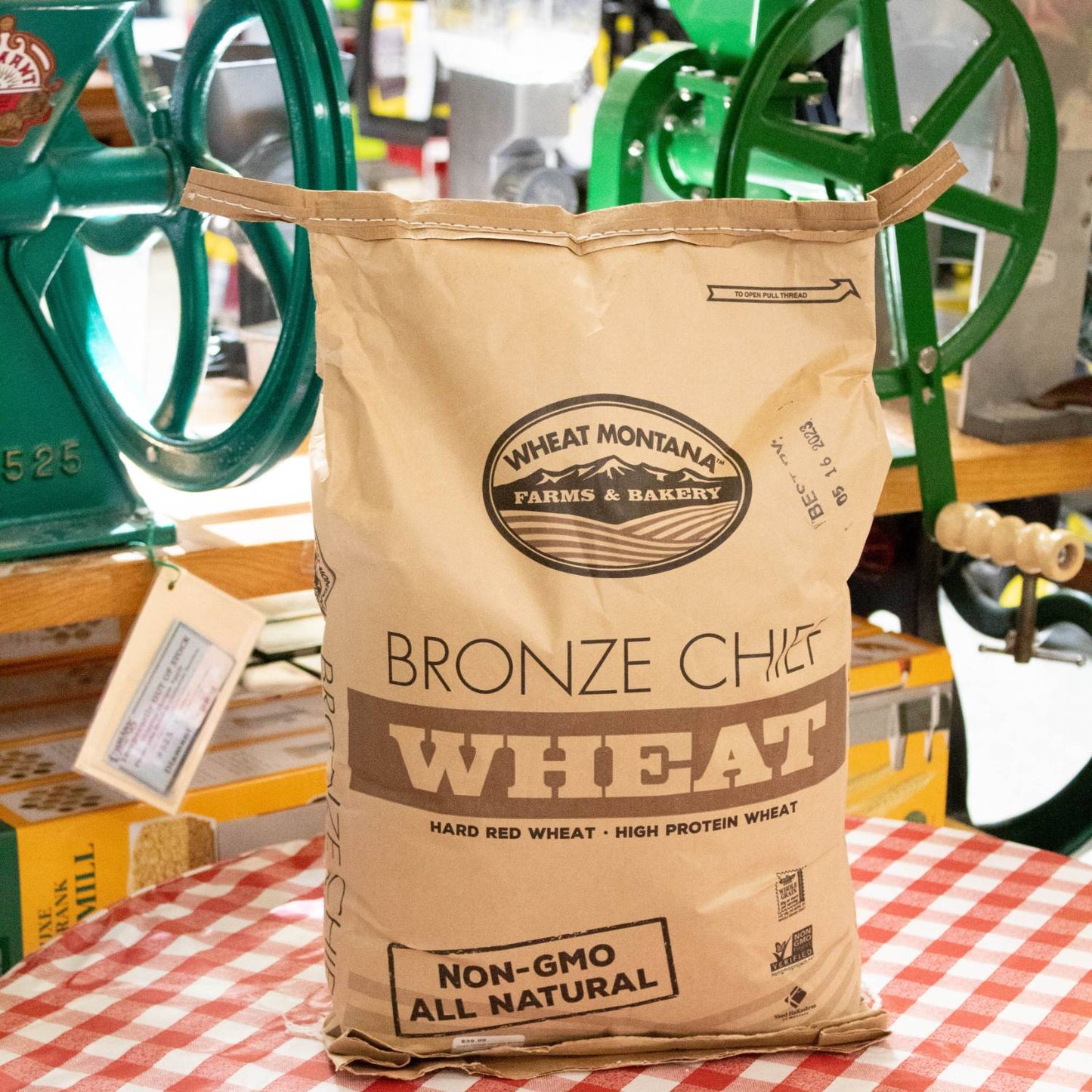 Large kraft paper bag of hard red wheat berries labeled non-GMO and all natural, placed on a checkered tablecloth with vintage grain mills in the background, ideal for Kitchen & Food and Bread Making Supplies collections.