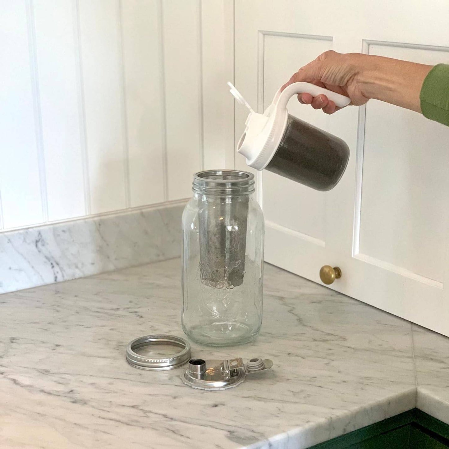 Hand placing a white mesh infuser filled with coffee grounds into a clear wide mouth jar on a marble countertop, showcasing a cold brew coffee maker accessory in a kitchen setting.
