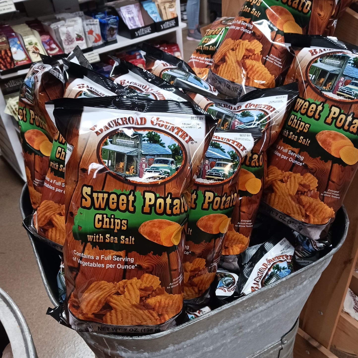 Bags of handcrafted sweet potato chips with sea salt, gluten free and made in USA, displayed in a rustic metal tub within Kitchen & Food and Snacks collections.