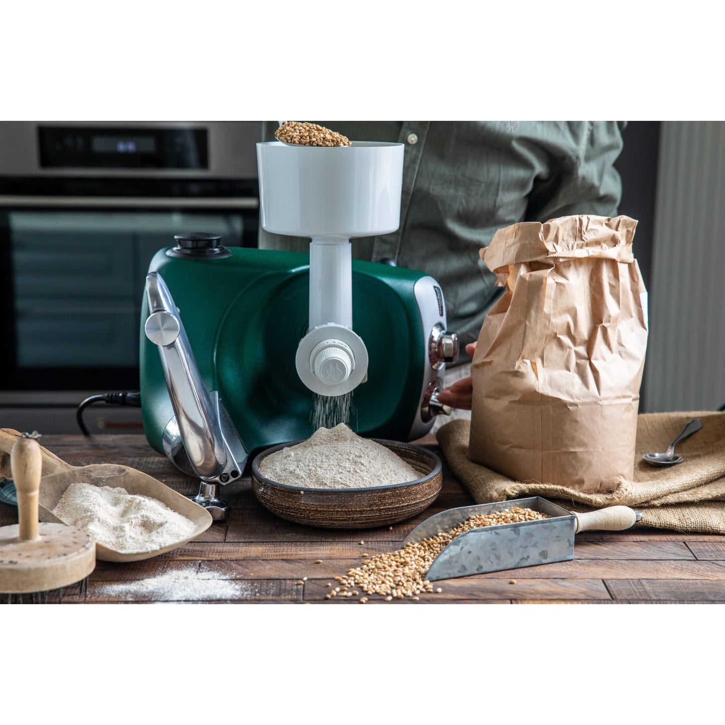 Green stand mixer fitted with a white grain mill attachment grinding fresh grain into flour collected in a bowl on a rustic wooden table with scattered wheat and a paper bag, ideal for Kitchen & Food, Baking Supplies, Grain Mills, and Bread Making collections.