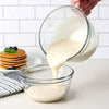 Clear thick glass batter bowl with spout and handle pouring pancake batter into matching bowl, set on kitchen counter with stack of pancakes nearby, ideal for Kitchen & Food and Baking Supplies collections.