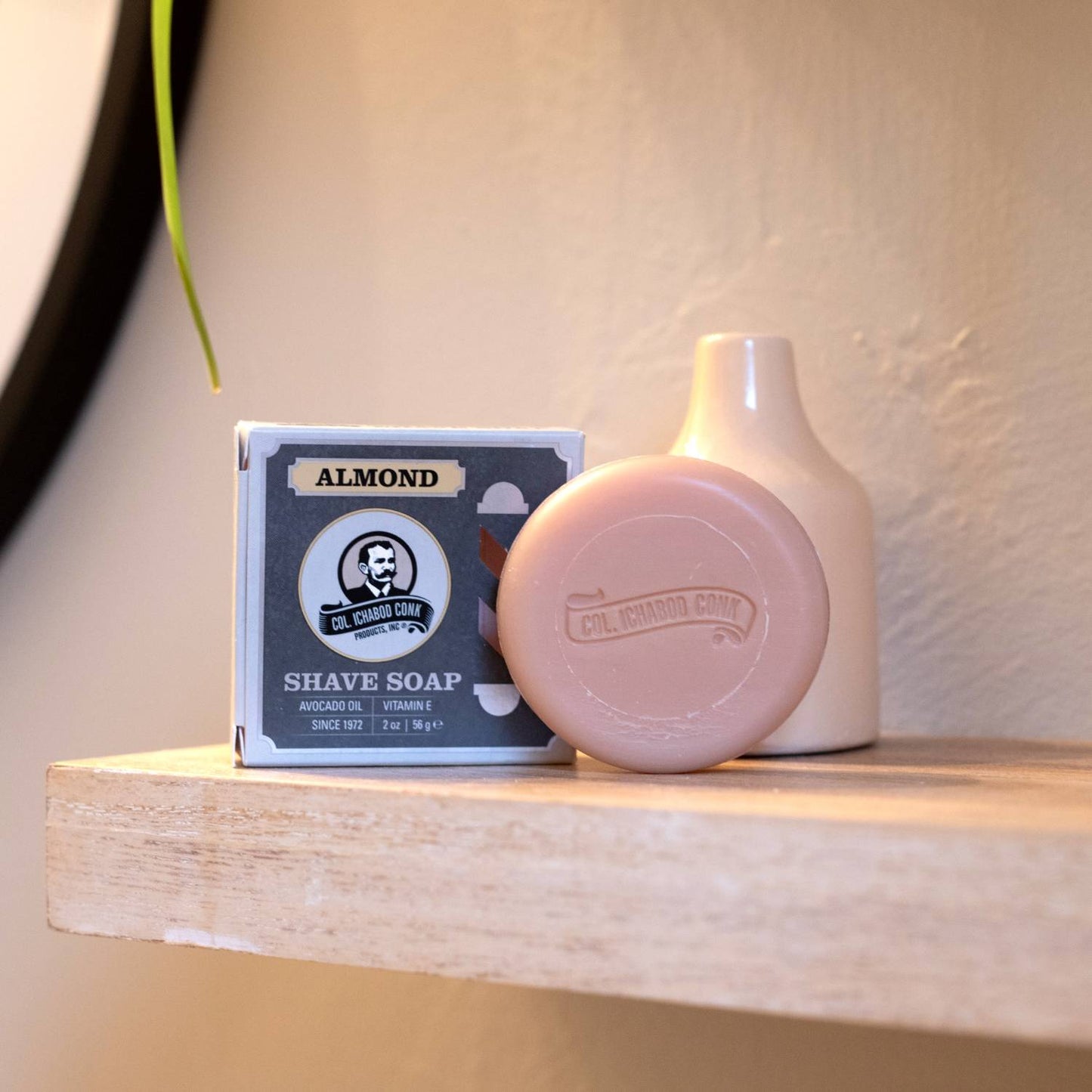 Round almond-colored shaving soap bar with embossed branding, displayed next to a matching box and minimalist white vase on a wooden shelf, featured in Health & Wellness, Grooming and Personal Hygiene, Soaps and Soapmaking, USA Made, Best Sellers, Promo Eligible collections.