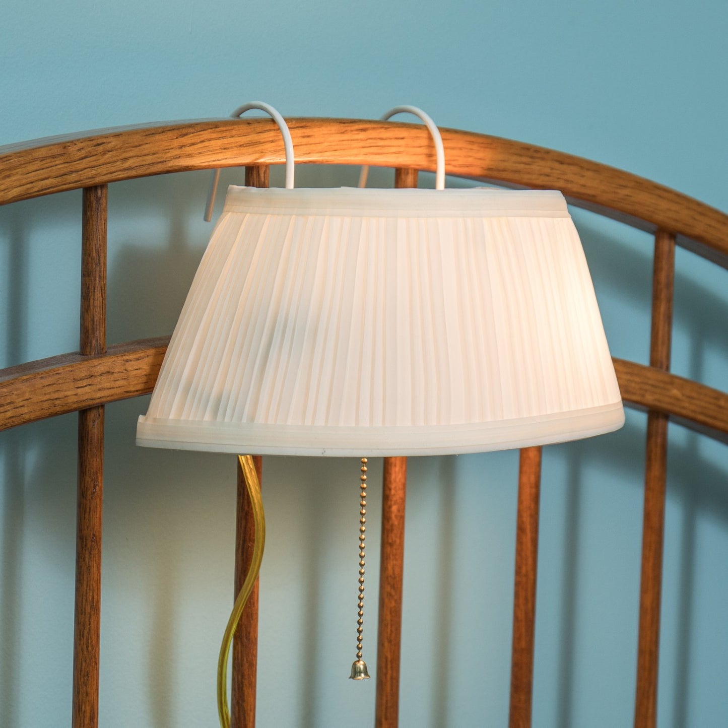 Soft white fabric lamp shade with gentle pleats hanging from curved hooks on a natural wood headboard, featuring a pull chain switch for convenient bedside lighting.