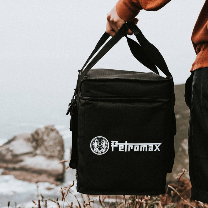 Person holding a black reinforced transport bag with shoulder strap, designed for outdoor stove carrying, featuring water-repellent ripstop fabric, ideal for camping and farm use.