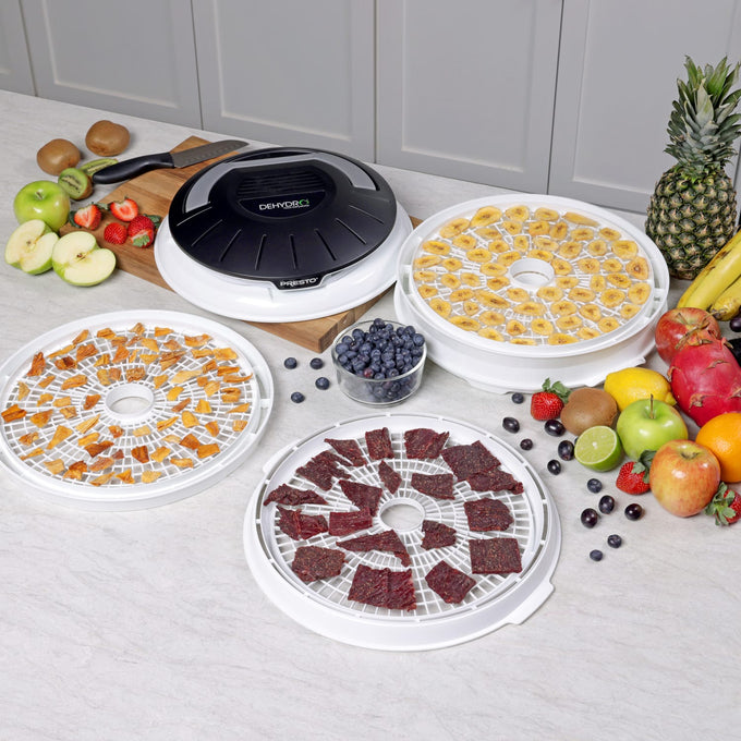 Compact electric food dehydrator with nesting white trays drying banana slices, beef jerky, and diced fruit, surrounded by fresh produce, suitable for Kitchen & Food and Food Preservation collections.