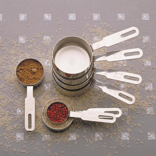 Set of heavy stainless steel measuring cups with clearly marked handles, nested and filled with brown sugar and red peppercorns, displayed on a kitchen surface with scattered rice grains in food prep collections.