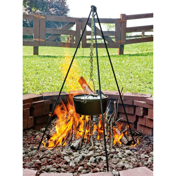 Tall Boy steel tripod with galvanized chain suspending a cast iron Dutch oven over an open campfire in a rustic outdoor stone fire pit, perfect for camping and outdoor cooking collections.