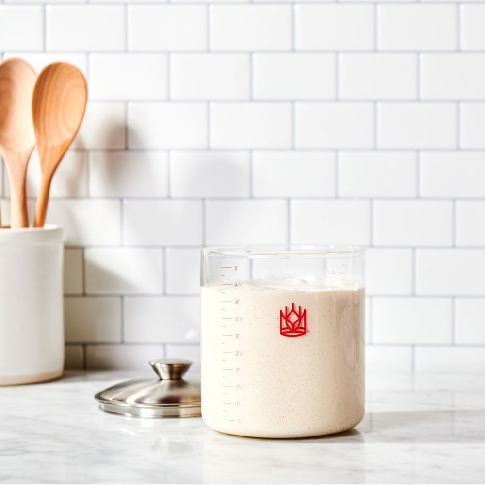 Clear glass sourdough crock filled with bubbly starter, accompanied by a stainless steel lid and wooden spoons in a ceramic holder, ideal for Kitchen & Food, Cookware, Baking Supplies, and Storage collections.