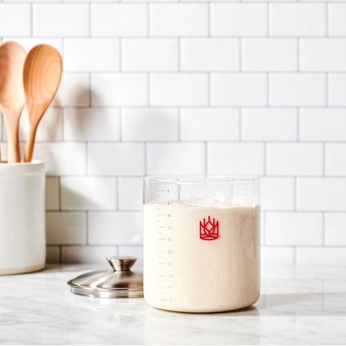 Clear glass sourdough crock filled with bubbly starter, accompanied by a stainless steel lid and wooden spoons in a ceramic holder, ideal for Kitchen & Food, Cookware, Baking Supplies, and Storage collections.