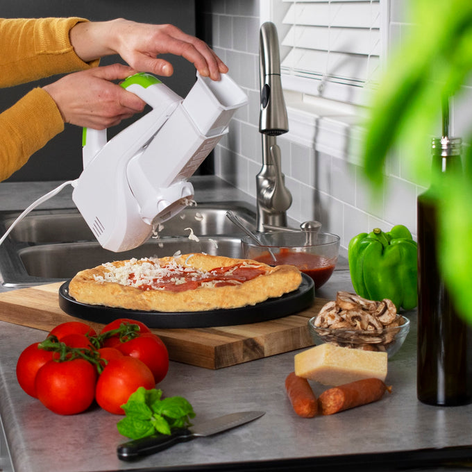 White and green electric kitchen gadget shredding cheese onto pizza crust on a cutting board with fresh tomatoes, green pepper, mushrooms, cheese block, and sausage nearby in a modern kitchen setting.