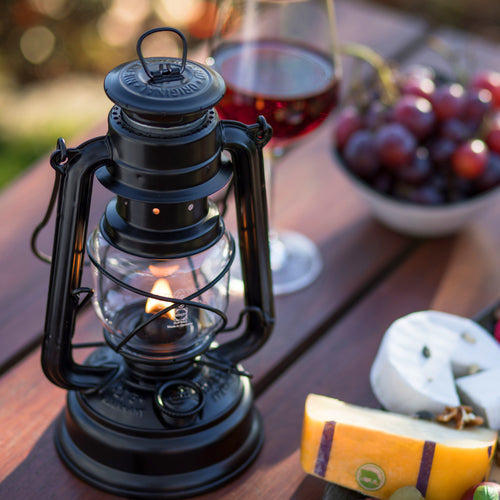 Handcrafted black galvanized oil lantern with thermal heat-resistant SUPRAX globe, glowing warmly on a wooden outdoor table with cheese, wine, and grapes, perfect for camping and emergency supplies in lamps & lighting collections.