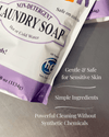 Partial view of a lavender and white package of non-detergent laundry soap featuring benefits for sensitive skin, simple natural ingredients, and powerful cleaning without synthetic chemicals, ideal for laundry supplies and soapmaking collections.