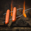 Chrome-plated dual-prong roasting fork with stay-cool wooden handle holding two hot dogs over a glowing campfire flame, perfect for camping and outdoor cooking.