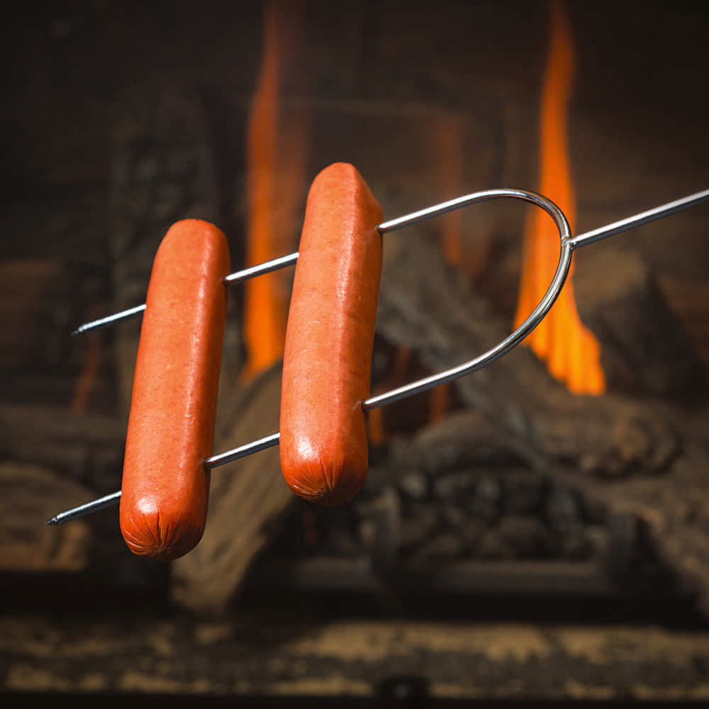 Chrome-plated dual-prong roasting fork with stay-cool wooden handle holding two hot dogs over a glowing campfire flame, perfect for camping and outdoor cooking.