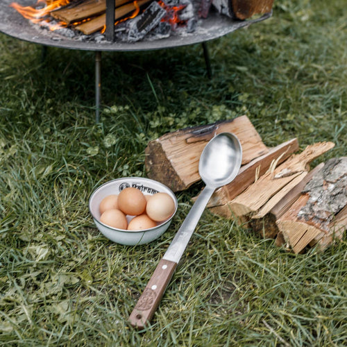 Handcrafted stainless steel serving spoon with a long walnut handle resting on grass near firewood and a bowl of eggs, perfect for Farm, Garden & Tool, Kitchen & Food, Camping, Storage and Serving, Best Sellers, Promo Eligible collections.
