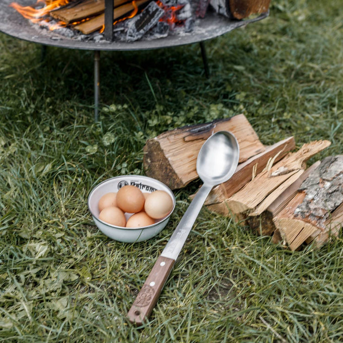 Handcrafted stainless steel serving spoon with a long walnut handle resting on grass near firewood and a bowl of eggs, perfect for Farm, Garden & Tool, Kitchen & Food, Camping, Storage and Serving, Best Sellers, Promo Eligible collections.