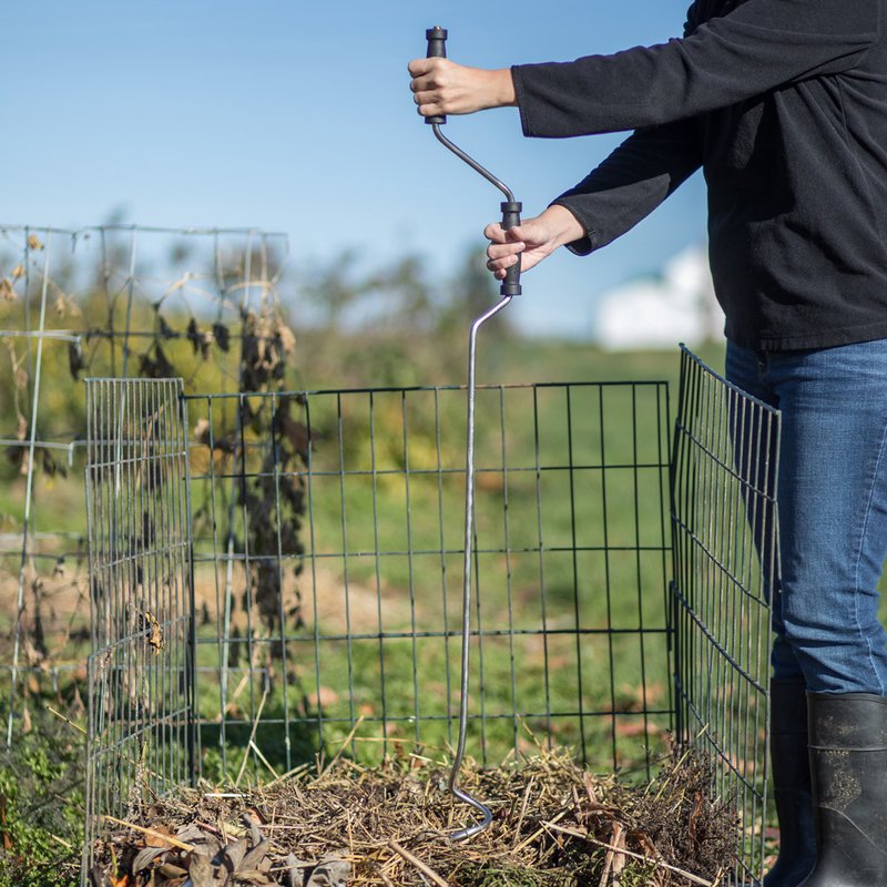 Person using a lightweight compost crank with a stainless steel spiral to mix organic material inside a metal garden compost bin, outdoors in a sunny farm or garden setting