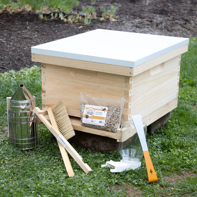Handcrafted natural wood beehive kit with essential beekeeping tools including smoker, brush, feeder, and hive tool, set on grass outdoors for farm, garden, and beekeeping supplies collections.