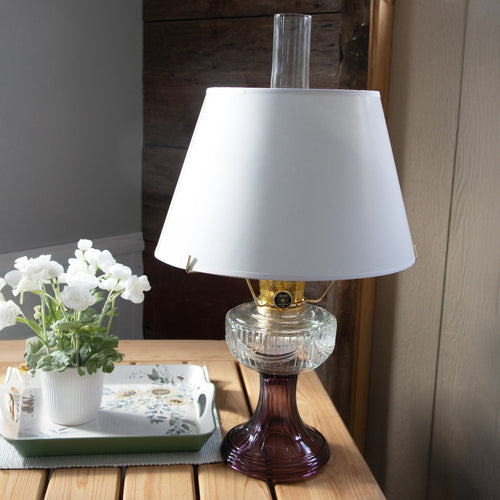White parchment oil lamp shade with tapered design atop a vintage-style glass oil lamp, displayed on a wooden table next to a potted white flower, fitting Lamps & Lighting and Parts and Accessories collections.