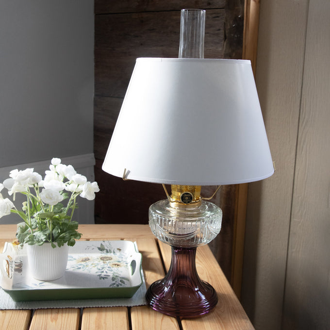 White parchment oil lamp shade with tapered design atop a vintage-style glass oil lamp, displayed on a wooden table next to a potted white flower, fitting Lamps & Lighting and Parts and Accessories collections.