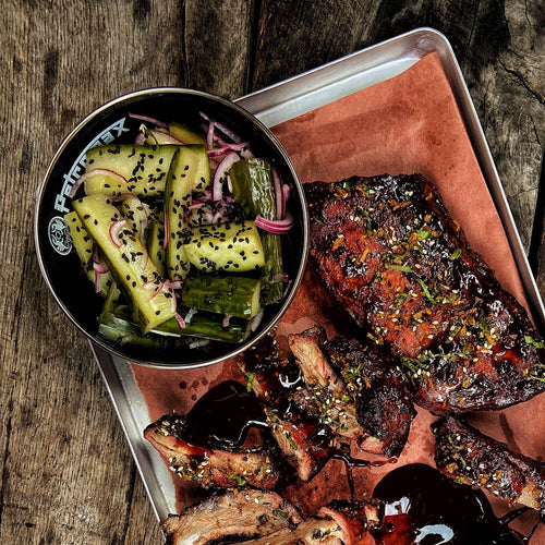 Black enamel bowl with white logo filled with seasoned pickles and onions, placed on a metal tray with grilled ribs, perfect for Farm, Garden & Tool, Kitchen & Food, Camping, Dinnerware, Best Sellers, and Promo Eligible collections.