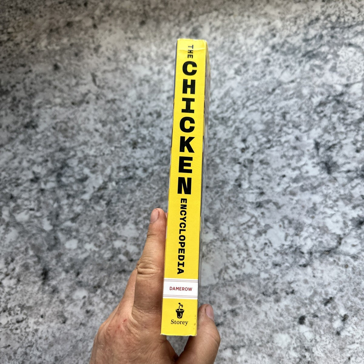 Hand holding the spine of a brightly colored yellow book titled with bold black text, part of Books, Farm and Livestock Books, Chicken Accessories, and Best Sellers collections.