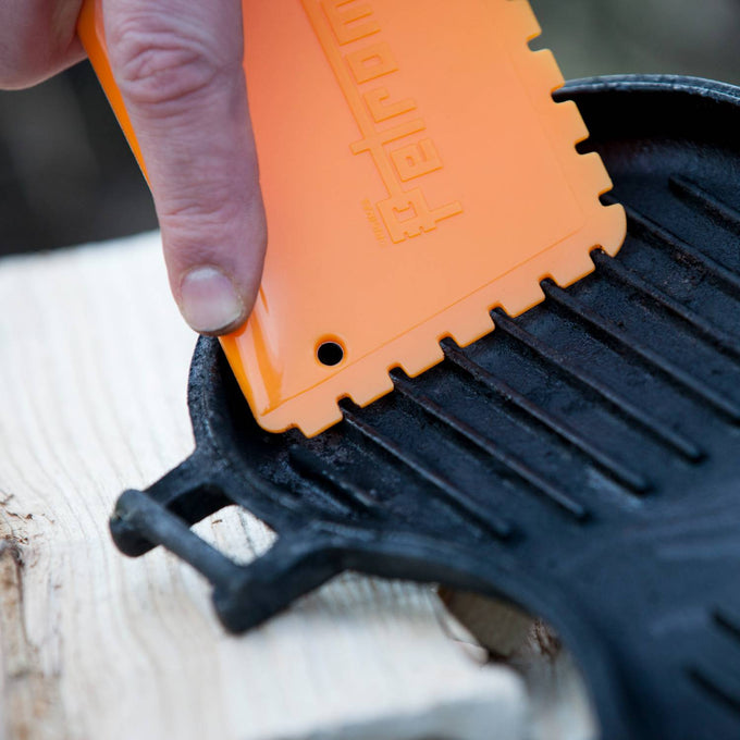 Hand using bright orange pan scraper gently cleaning residue from a cast iron grill pan, designed to protect seasoning while restoring cookware, ideal for Kitchen & Food and Cast Iron collections.