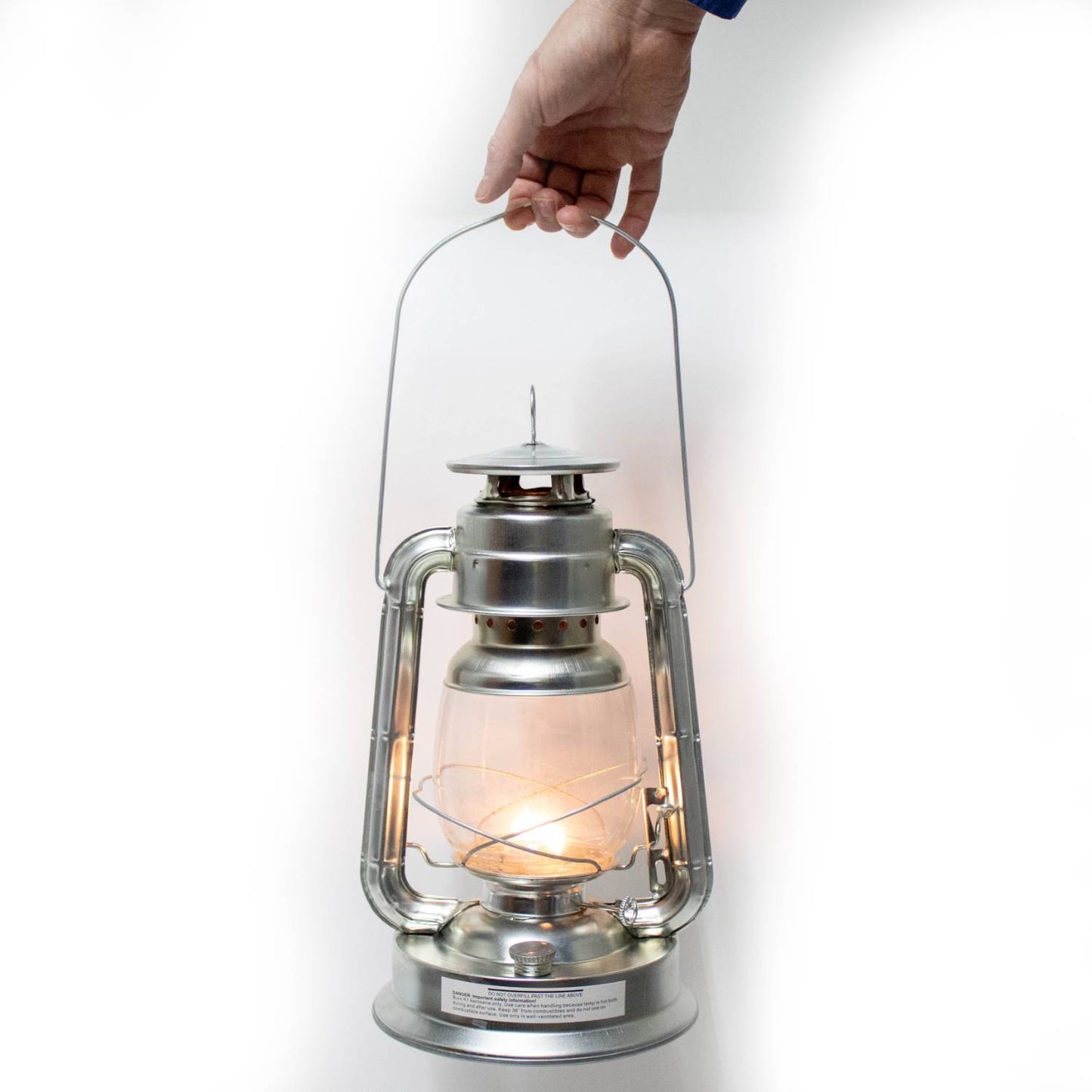 Hand holding a silver metal oil lantern with lit flame inside clear glass globe, designed for outdoor use in camping, pest control, and emergency supplies collections.