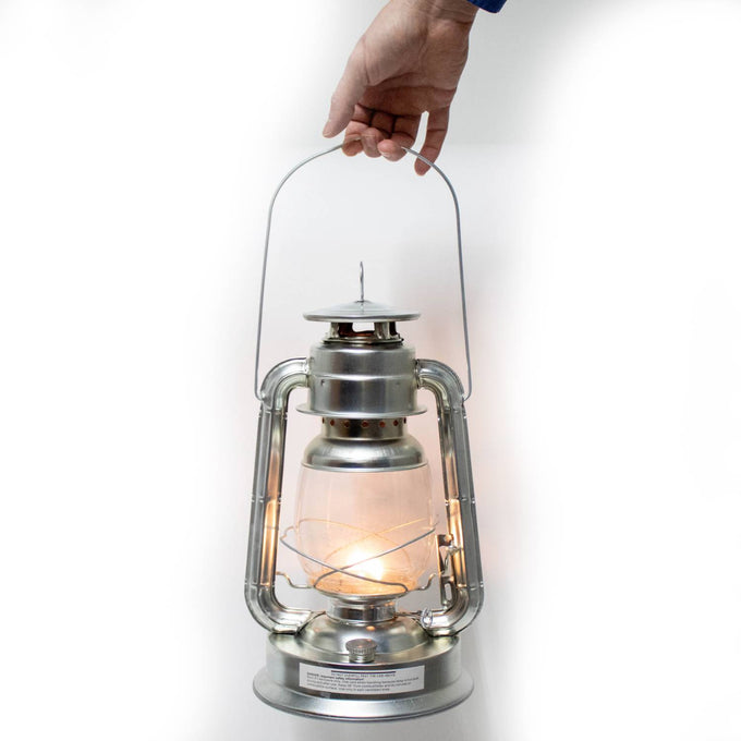 Hand holding a silver metal oil lantern with lit flame inside clear glass globe, designed for outdoor use in camping, pest control, and emergency supplies collections.