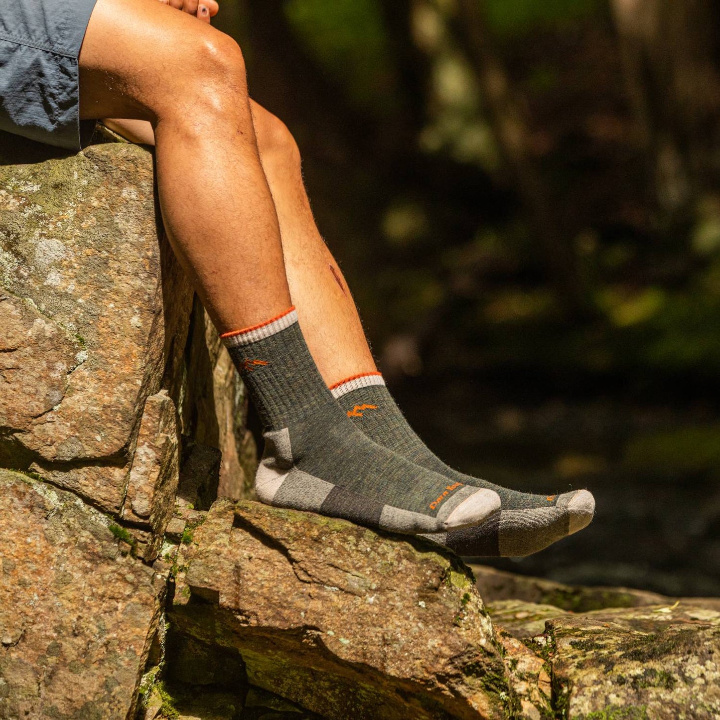 Man sitting on natural rock formation wearing cushioned midweight hiking socks with reinforced heel and toe, crafted in USA, ideal for camping, outdoor activities, and best sellers at Lehman’s.