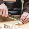 Person using a handcrafted utility knife with a high-quality steel blade and walnut handle to slice fresh mushrooms on a wooden cutting board in a kitchen setting, ideal for food prep and camping.