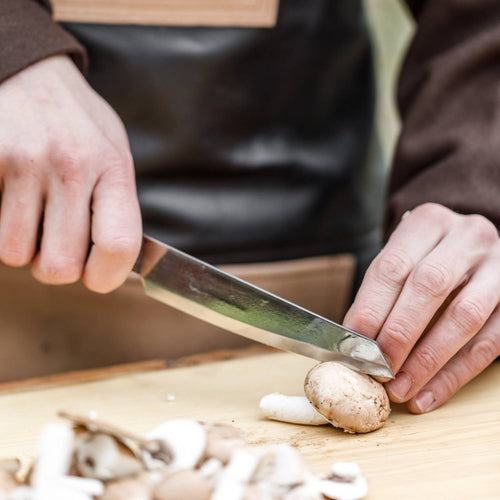 Person using a handcrafted utility knife with a high-quality steel blade and walnut handle to slice fresh mushrooms on a wooden cutting board in a kitchen setting, ideal for food prep and camping.