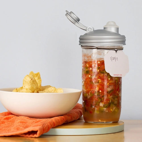 Clear wide-mouth jar with a silver fermentation lid and airlock filled with mixed vegetable ferment, labeled with a handwritten date tag, displayed next to a bowl of potato chips on an orange cloth, ideal for Kitchen & Food, Food Preservation, Fermenting and Pickling, USA Made, and Best Sellers collections.