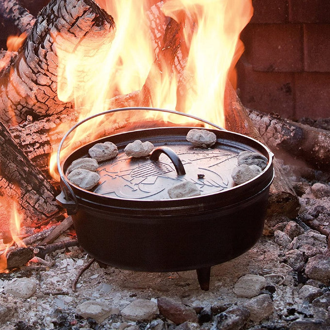 Cast iron camp Dutch oven with legs cooking over an open fire surrounded by glowing embers, featuring a handle and rocks on the lid, ideal for camping and outdoor cookware collections.