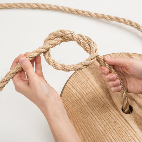 Close-up of hands tying a sturdy natural fiber rope through the center hole of a round wooden swing seat, handcrafted and made in USA, ideal for outdoor play and children’s toys collections.