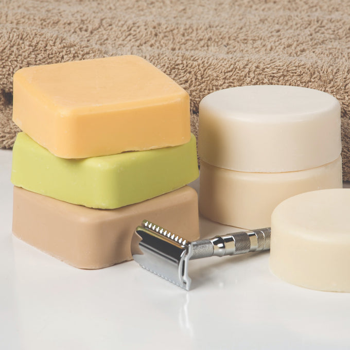 Three handmade square soap bars in orange, green, and tan colors stacked near round cream-colored bars and a metal safety razor, featured in Health & Wellness, Grooming and Personal Hygiene, Soaps and Soapmaking, Shampoo and Conditioner, USA Made, and Best Sellers collections.