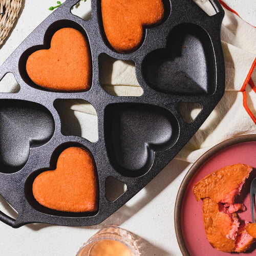 Cast iron mini heart-shaped cake pan with several baked orange-colored heart cakes, shown with one partially eaten cake on a pink plate, ideal for Kitchen & Food, Cookware, Cast Iron, Baking Supplies, USA Made collections.