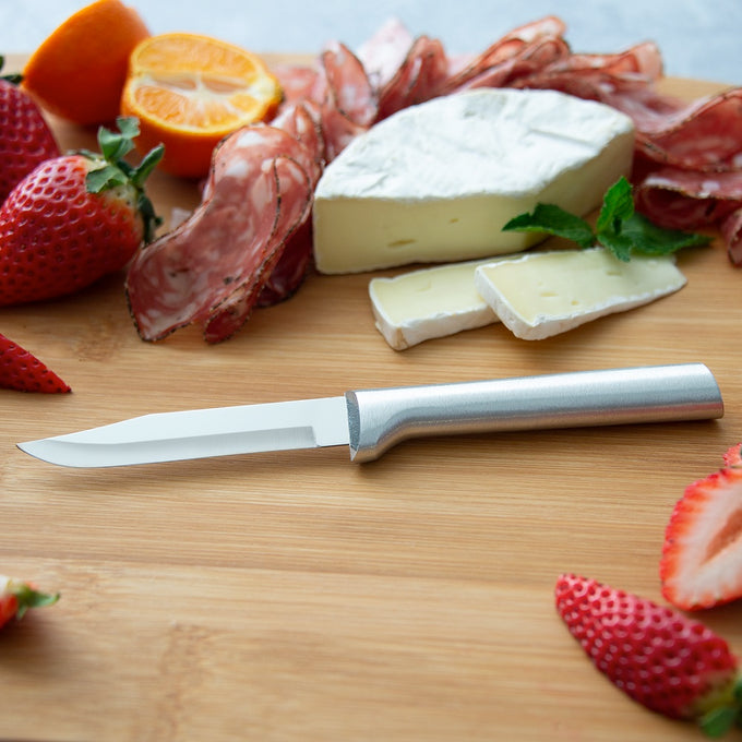 Sharp carbon stainless steel paring knife with a satin-finished cast aluminum handle, displayed on a wooden board with sliced strawberries, orange, cheese, and cured meats in a kitchen and food prep setting.