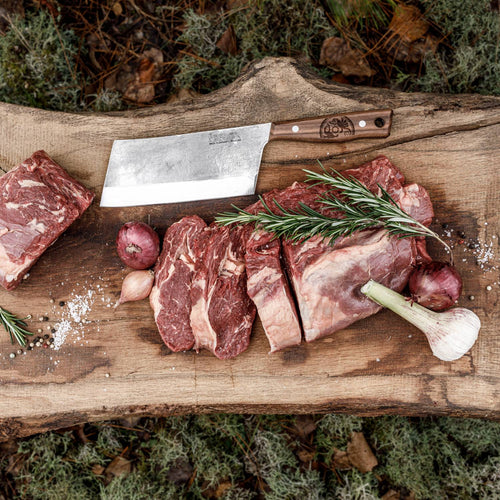 Handcrafted cleaver knife with a sturdy hardened blade and wooden handle resting on a rustic wooden board next to fresh sliced meat, garlic, shallots, and rosemary, suited for butchering, camping, and kitchen food prep in Farm, Garden & Tool and Specialty Equipment collections.