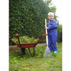 Child in blue coveralls using a small garden rake near a red wheelbarrow filled with green garden clippings, set against a neatly trimmed hedge in a lawncare and garden accessories setting.