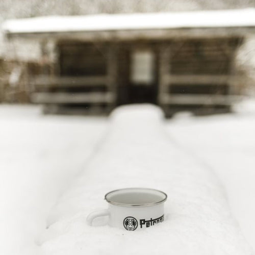 White enamel steel mug with metal rim and black logo placed on a snow-covered surface in front of a rustic wooden cabin, suited for Farm, Garden & Tool, Camping, Brewing Coffee and Tea, Dinnerware, Best Sellers, and Promo Eligible collections.