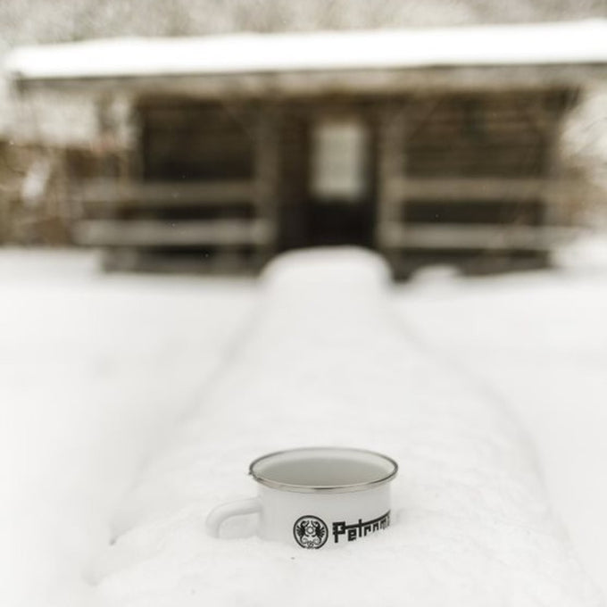 White enamel steel mug with metal rim and black logo placed on a snow-covered surface in front of a rustic wooden cabin, suited for Farm, Garden & Tool, Camping, Brewing Coffee and Tea, Dinnerware, Best Sellers, and Promo Eligible collections.
